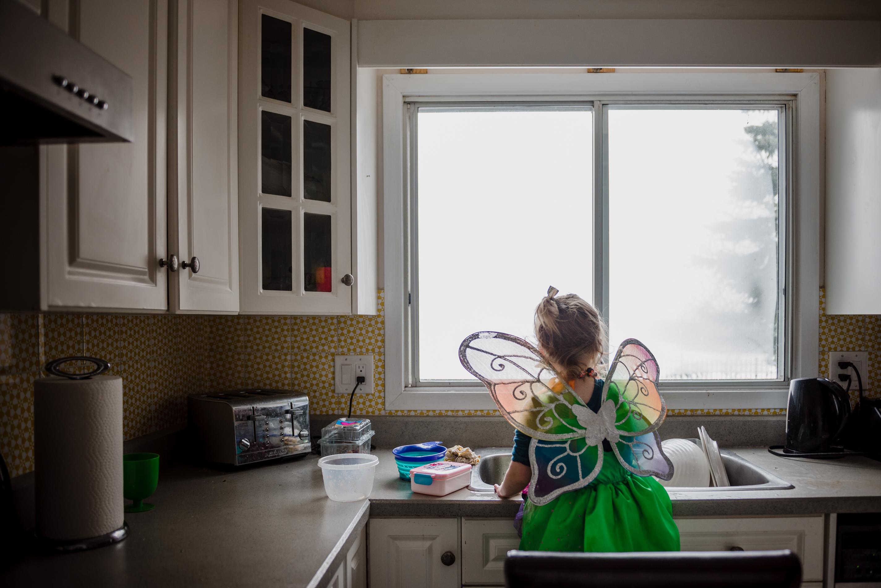 A toddler girl is dressed as Tinkerbell and a butterfly washing dishes.