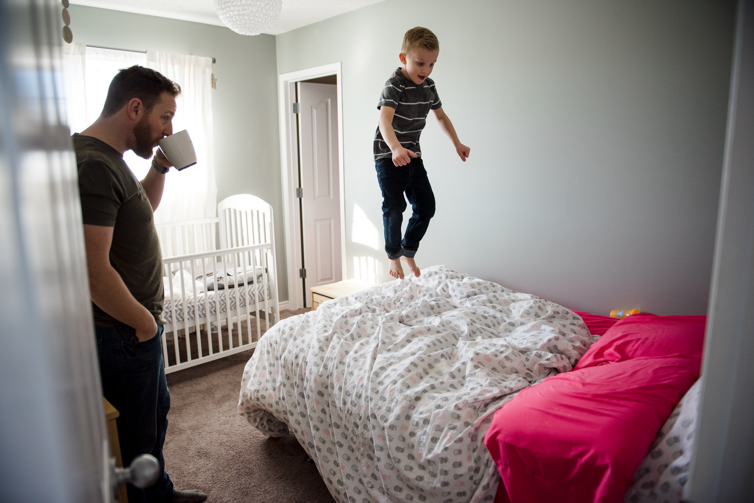 A dad drinks coffee while watching his son jump on the bed in his bedroom