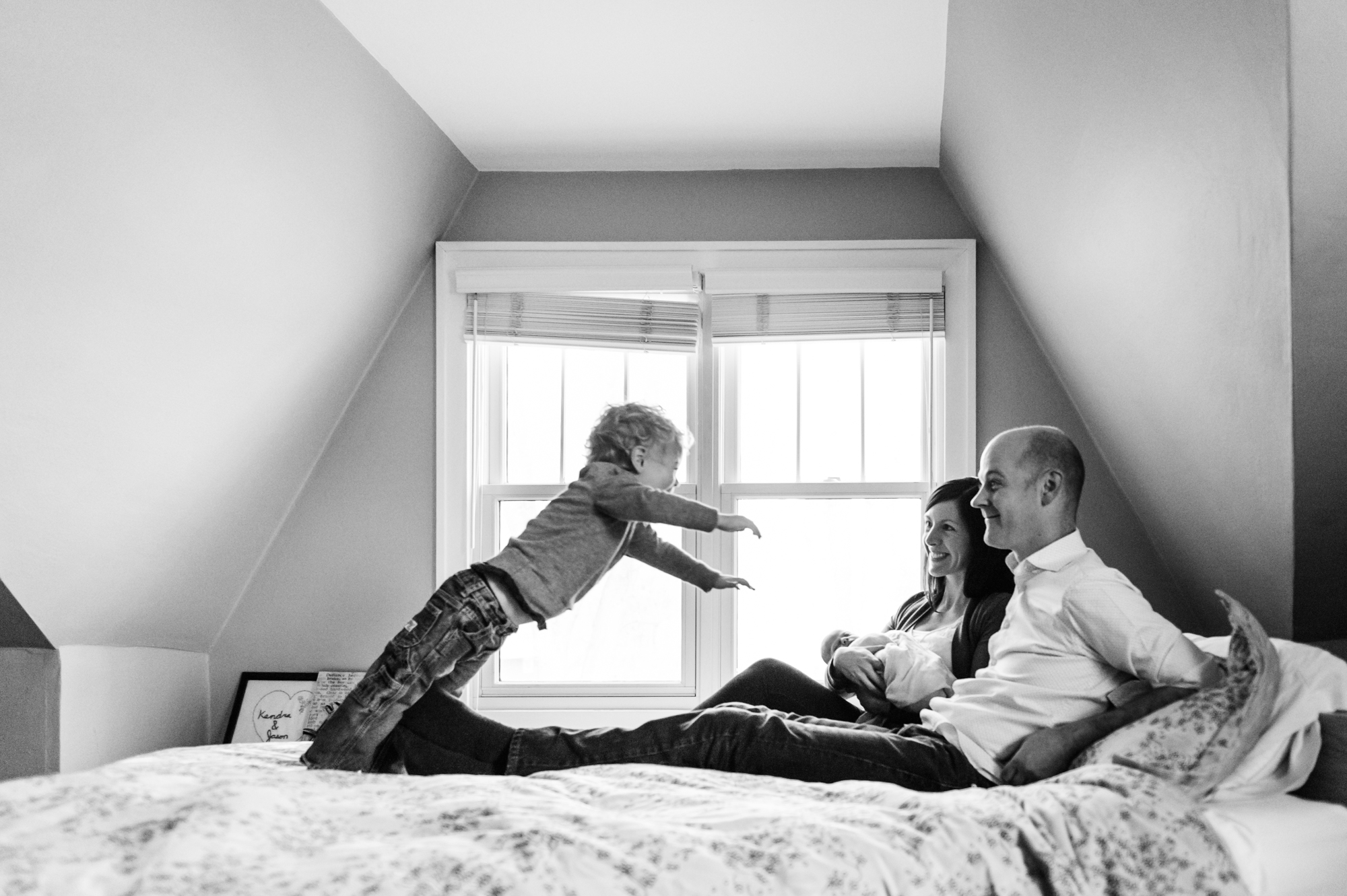 A family sits on a bed during their newborn photos session while big brother jumps