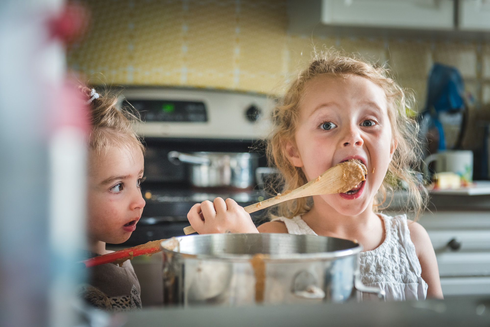 A little sister watches on as her big sister licks batter from a spoon