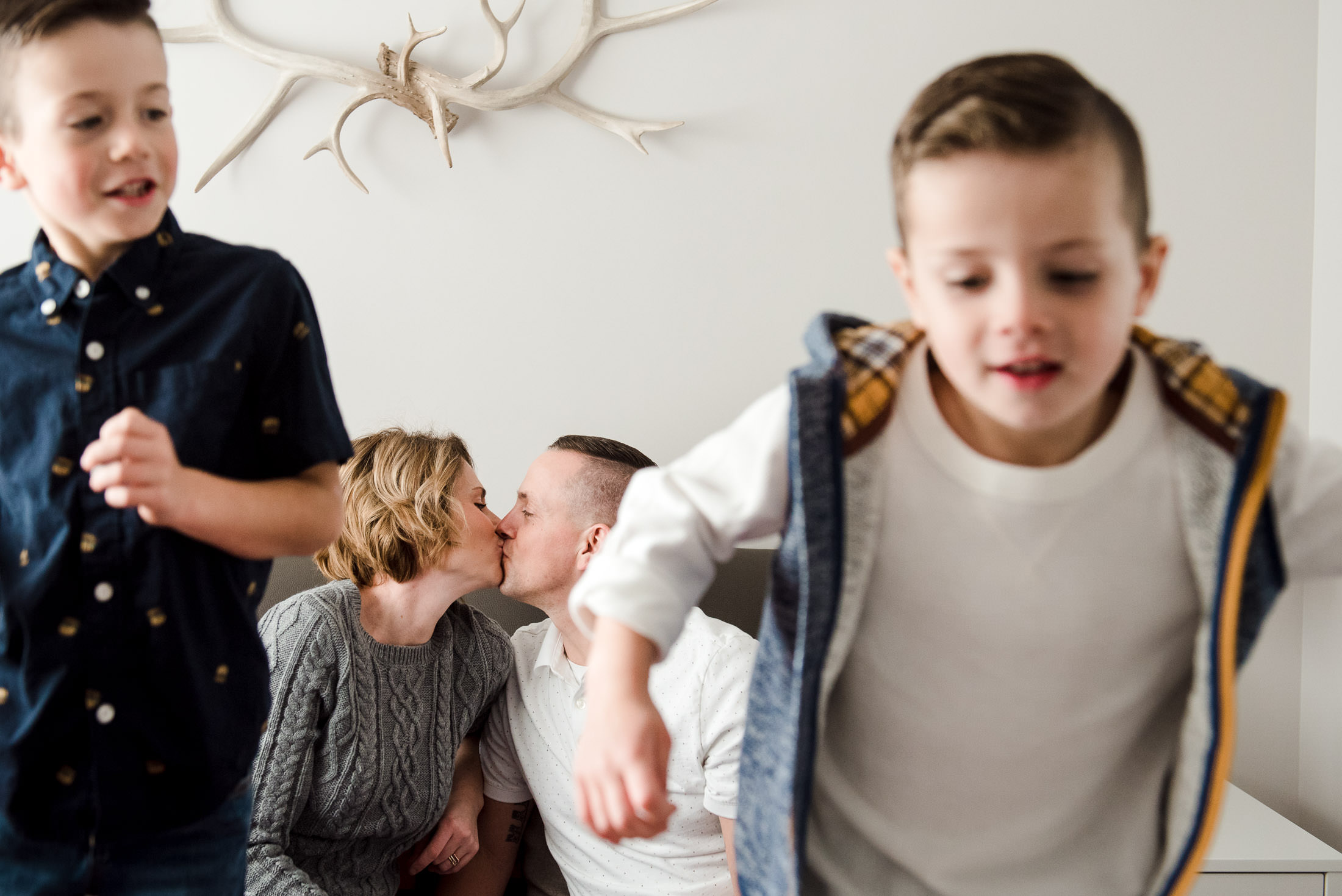 A mom and dad kiss while the kids jump on the bed during family photos