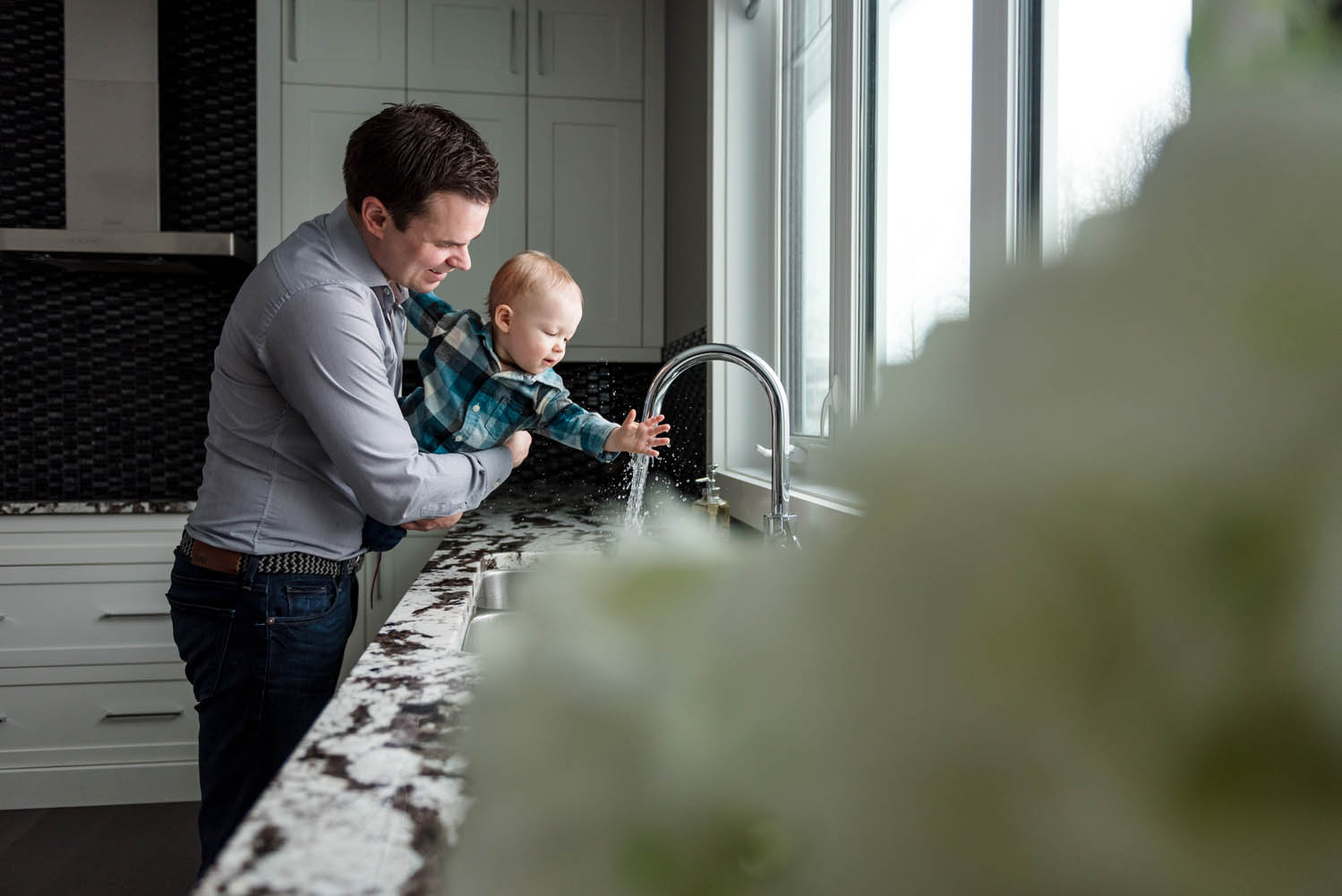 A dad splashes in the kitchen sink with his 1 year old baby during a family photo session.