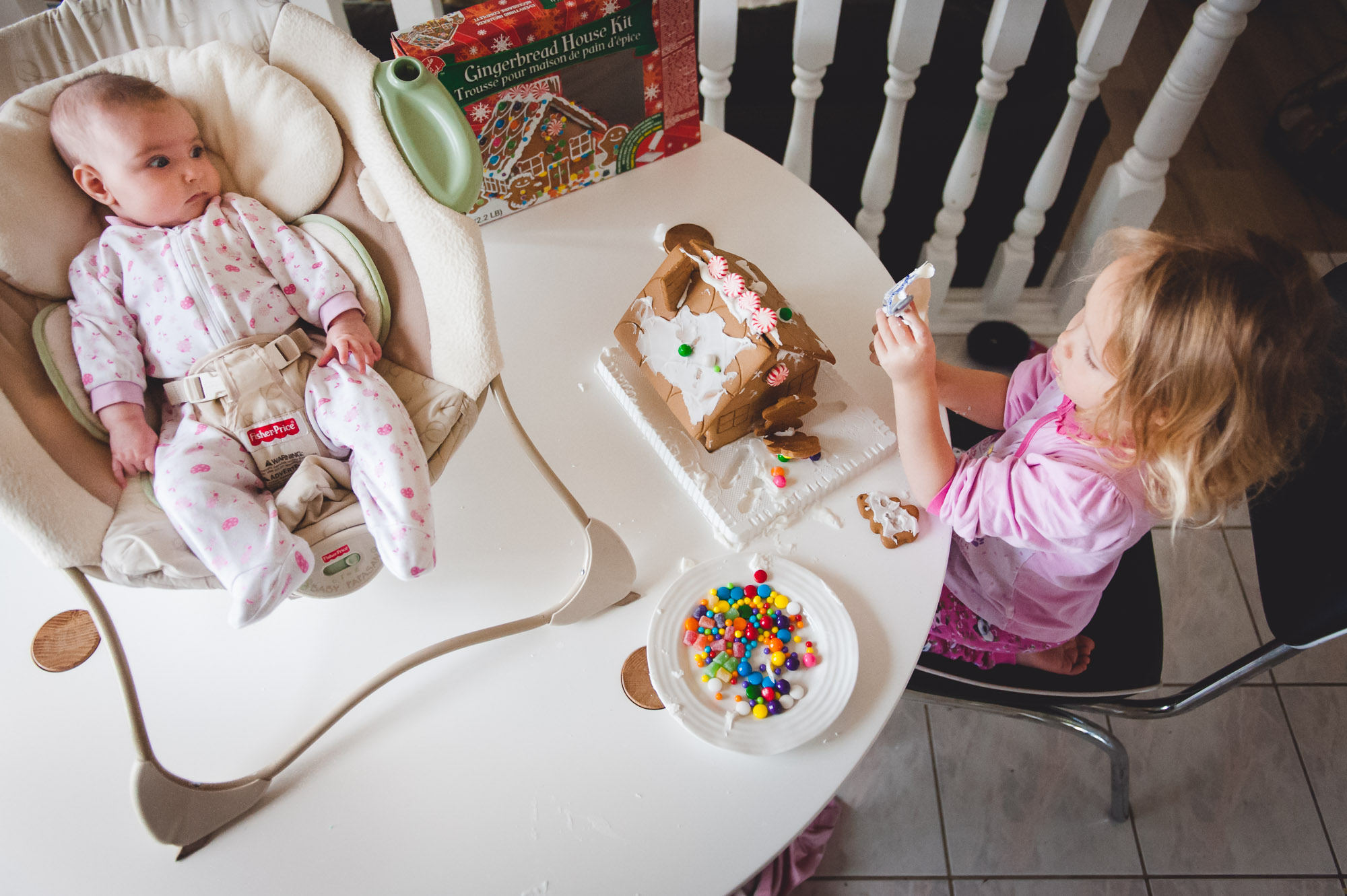 A baby sits by and watches her big sister build a gingerbread house during a documentary photo session