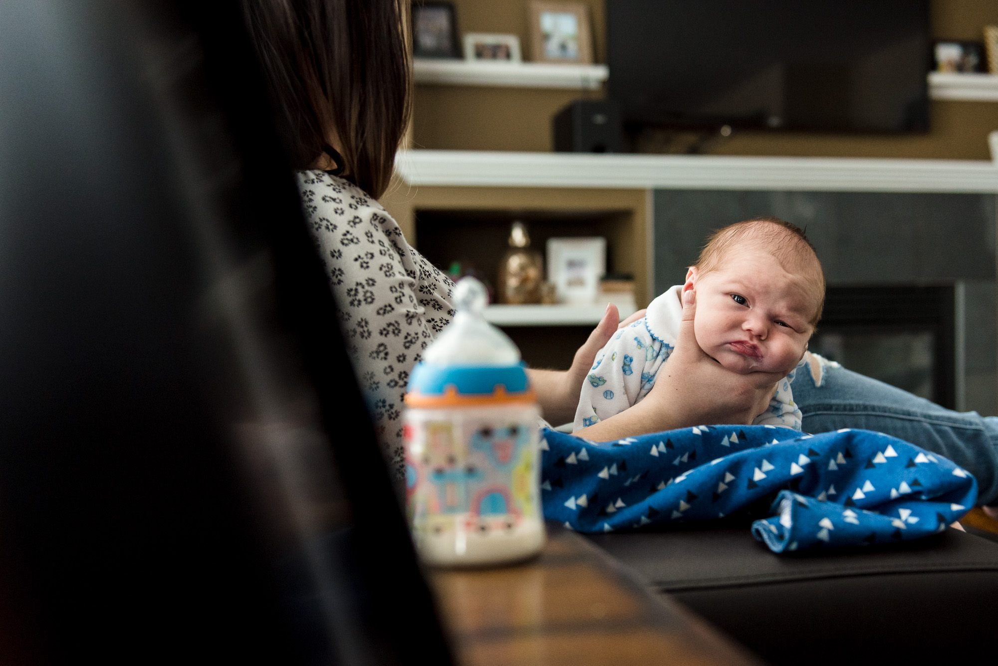A mom burps a newborn baby in their Sherwood Park living room while the empty bottle sits nearby. 