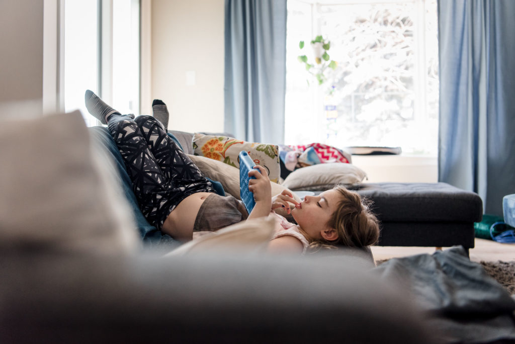 A girl plays her tablet while hanging upside down on the couch in her Edmonton home. 