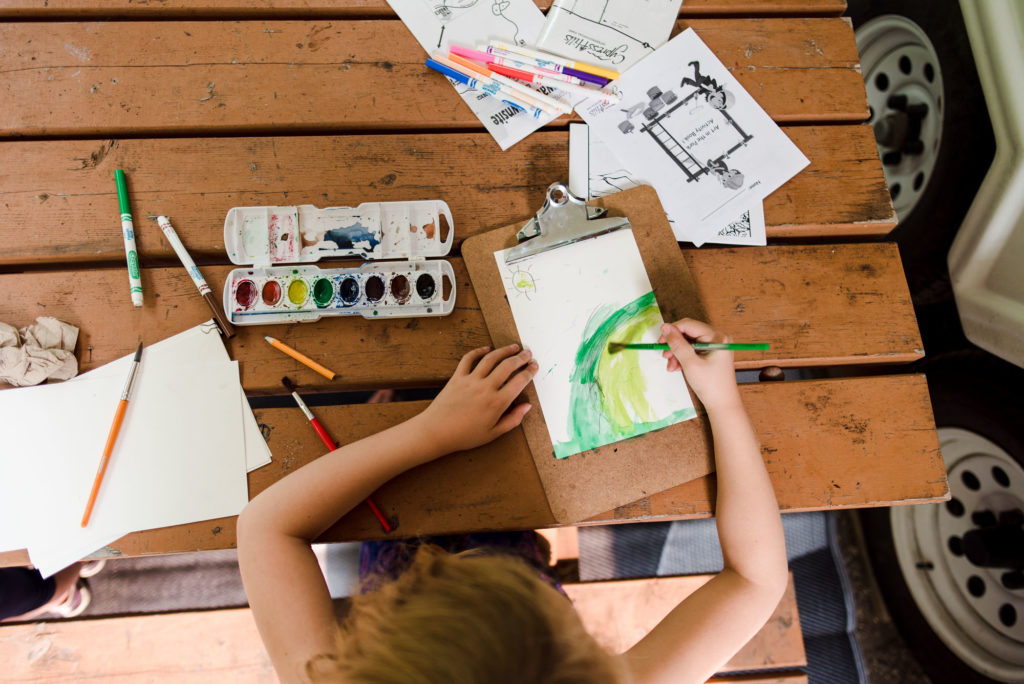 A girl paints watercolours while at a picnic table. Things to bring with you with camping to entertain kids
